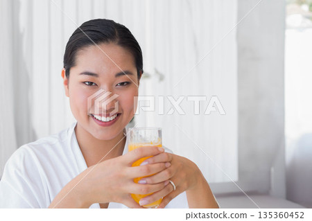 Korean woman sitting in bright home lounge wearing white robe, holding clear glass of orange juice Korean woman sitting in bright home lounge wearing white robe, holding clear glass of orange juice 135360452
