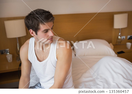 Man sitting on edge of unmade bed in hotel-style bedroom wearing pajamas checking smartphone Man sitting on edge of unmade bed in hotel-style bedroom wearing pajamas checking smartphone 135360457