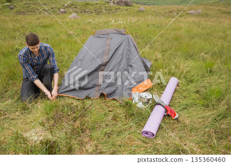 Man kneeling on grassy meadow securing tent stake beside pitched tent by purple mat, copy space 135360460