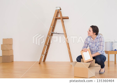 Woman in mid-20s crouching near ladder unpacking picture frame at home with paint cans, copy space Woman in mid-20s crouching near ladder unpacking picture frame at home with paint cans, copy space 135360464