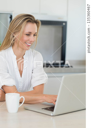 Middle-aged woman smiling and typing on laptop at kitchen countertop with ceramic mug, copy space Middle-aged woman smiling and typing on laptop at kitchen countertop with ceramic mug, copy space 135360467