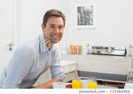 Mid-adult man sitting at kitchen table leaning on table smiling with orange juice cereal coffee cup Mid-adult man sitting at kitchen table leaning on table smiling with orange juice cereal coffee cup 135360468