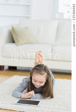 Girl lying prone on light-colored shag rug at home tapping tablet computer near white sofa Girl lying prone on light-colored shag rug at home tapping tablet computer near white sofa 135360491