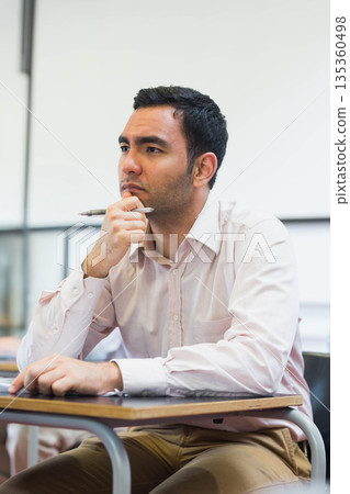 Male student sitting at desk at school leaning forward and holding pen near whiteboard 135360498