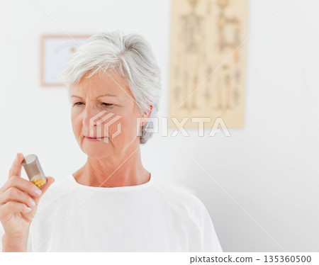 Senior female holding translucent pill bottle examining label in exam room with chart and document 135360500