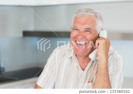 Senior man talking on corded telephone in modern kitchen with countertop and cabinets, copy space Senior man talking on corded telephone in modern kitchen with countertop and cabinets, copy space 135360502