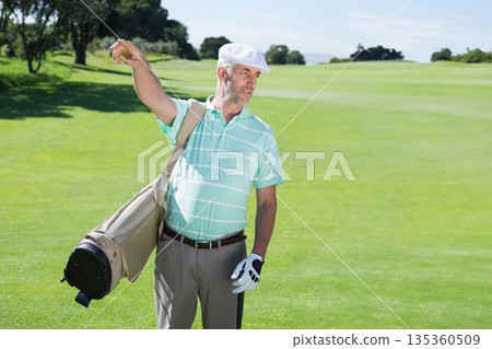 Male golfer pointing toward fairway on golf course while holding club and carrying golf bag Male golfer pointing toward fairway on golf course while holding club and carrying golf bag 135360509