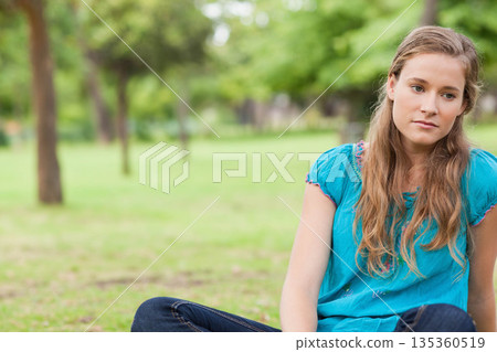 Woman in bright teal blouse and jeans sitting cross-legged on grassy field in park, copy space 135360519