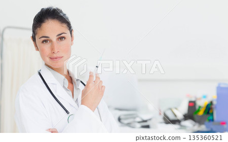 Female physician holding syringe upright in exam room wearing coat and stethoscope, copy space 135360521