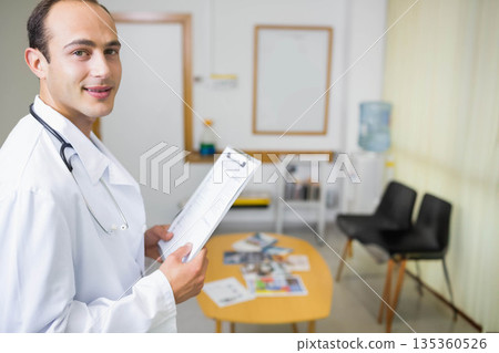 Hispanic adult male doctor in white lab coat with stethoscope standing in clinic holding clipboard Hispanic adult male doctor in white lab coat with stethoscope standing in clinic holding clipboard 135360526