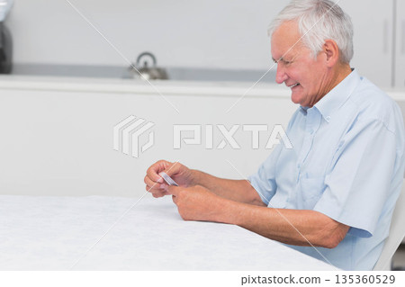Senior man holding playing cards and smiling at dining table in kitchen with kettle, copy space 135360529