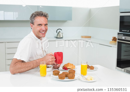 Middle-aged man smiling while holding red mug at home kitchen table with cereal and orange juice Middle-aged man smiling while holding red mug at home kitchen table with cereal and orange juice 135360531