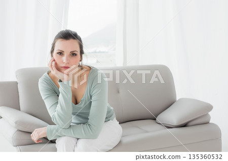 Woman in her thirties sitting on grey leather sofa in living room gazing through sheer curtains Woman in her thirties sitting on grey leather sofa in living room gazing through sheer curtains 135360532