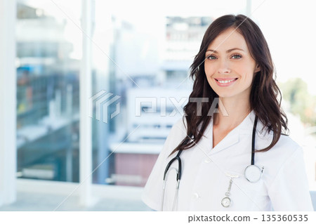 Female doctor standing in clinic office wearing white coat with stethoscope and badge, copy space 135360535