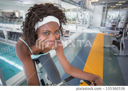 African American woman wearing green sports bra gripping handlebars pedaling exercise bike in gym 135360536
