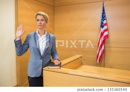 Mid adult woman raising hand, resting on book at witness stand with American flag, copy space Mid adult woman raising hand, resting on book at witness stand with American flag, copy space 135360549
