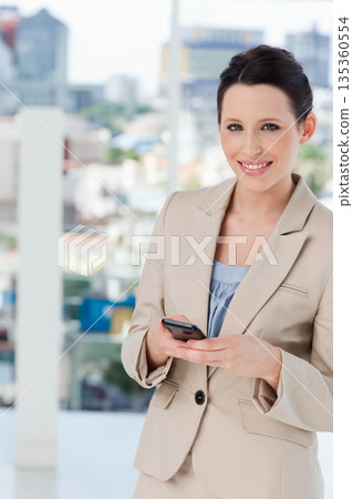 Businesswoman smiling while holding smartphone on balcony overlooking city skyline, copy space 135360554