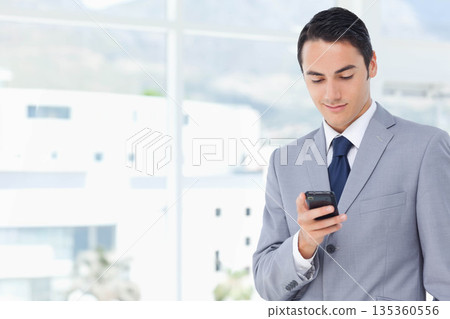 Male in business suit holding smartphone while checking emails in bright office space, copy space Male in business suit holding smartphone while checking emails in bright office space, copy space 135360556