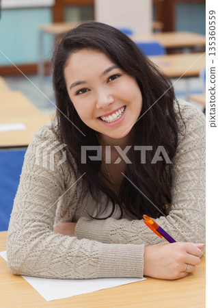 Asian woman smiling while writing on paper with purple-and-orange pen at wooden desk in classroom 135360559