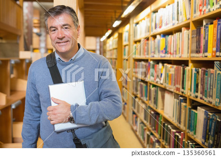 Male in his fifties standing in library aisle holding tablet computer with shoulder bag, copy space 135360561