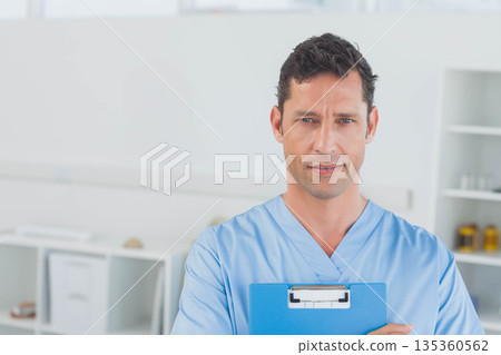 Mature adult man standing in clinical examination room holding blue clipboard with medical supplies 135360562