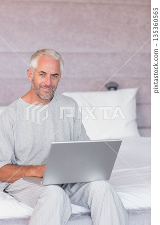 Senior man sitting on bed in softly lit bedroom holding silver laptop near padded headboard 135360565