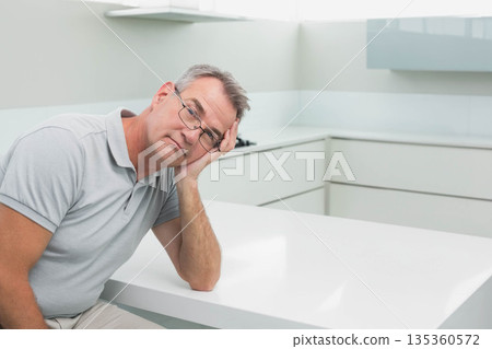 Middle-aged man wearing glasses leaning head on hand sitting at white kitchen island, copy space Middle-aged man wearing glasses leaning head on hand sitting at white kitchen island, copy space 135360572