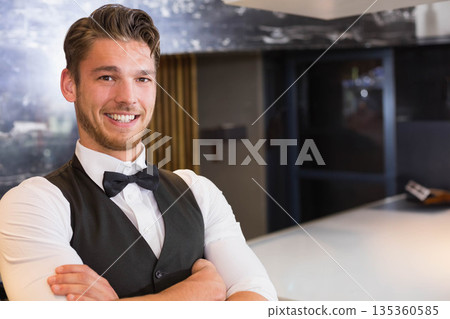 male bartender in black vest smiling and standing at lounge bar counter with bag, copy space 135360585