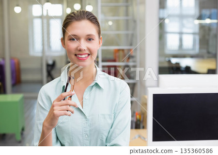 Woman wearing green shirt holding eyeglasses and smiling at camera at desk in office with monitor Woman wearing green shirt holding eyeglasses and smiling at camera at desk in office with monitor 135360586