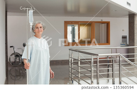 Senior female patient holding IV pole with saline bag in ward corridor near wheelchair, copy space 135360594