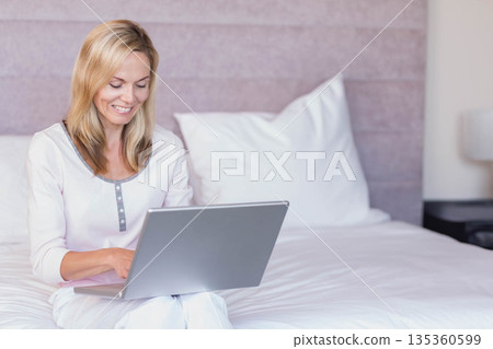 Middle-aged woman sitting cross-legged on bed typing on laptop in bedroom with pillow and lamp 135360599
