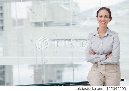 Woman sitting on glass desk in office crossing arms and smiling at window blinds, copy space 135360603