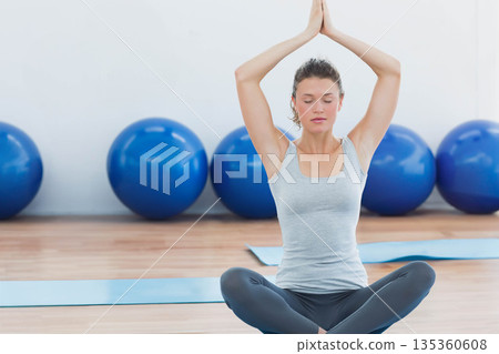 Woman sitting cross-legged on blue yoga mat in yoga studio with exercise balls behind in sportswear 135360608