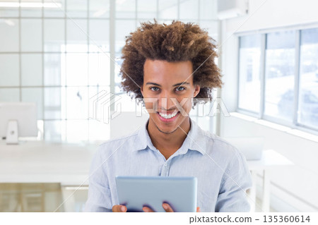 African American man wearing light blue shirt standing in modern office holding tablet and smiling 135360614