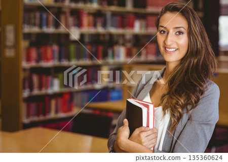 Middle Eastern woman standing at library with wooden tables holding two books smiling, copy space Middle Eastern woman standing at library with wooden tables holding two books smiling, copy space 135360624
