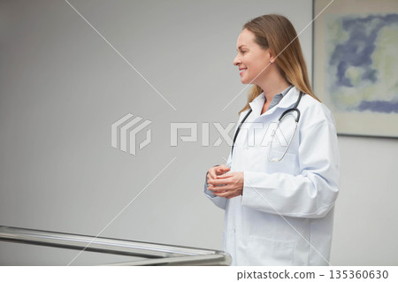 Woman doctor standing beside railing in clinic corridor in lab coat holding stethoscope, copy space Woman doctor standing beside railing in clinic corridor in lab coat holding stethoscope, copy space 135360630