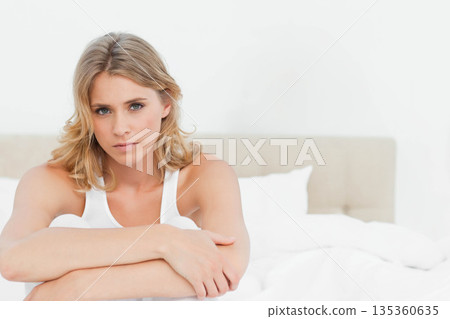 Adult woman hugging knees, looking forward on bed in bedroom with white linens and headboard 135360635