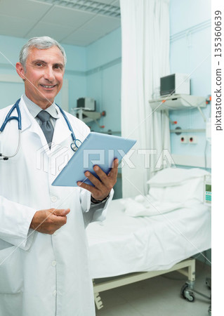 Senior male doctor standing at bedside holding tablet wearing lab coat and stethoscope, copy space Senior male doctor standing at bedside holding tablet wearing lab coat and stethoscope, copy space 135360639