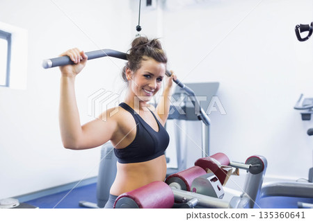 Woman smiling while pulling wide pulley bar on lat pulldown machine at gym during strength training Woman smiling while pulling wide pulley bar on lat pulldown machine at gym during strength training 135360641