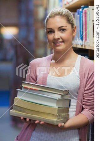 Female student standing in library aisle holding stack of textbooks near bookshelves 135360643