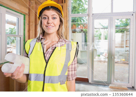 Woman wearing hard hat and vest, holding blueprints and reviewing plans inside renovation site 135360644