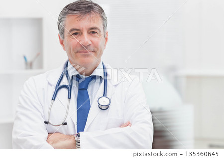 Senior male doctor standing with arms crossed in clinic, wearing white lab coat and stethoscope Senior male doctor standing with arms crossed in clinic, wearing white lab coat and stethoscope 135360645