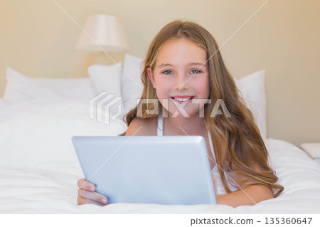 Child girl lying on bed in bedroom propped on elbows holding tablet among pillows near lamp Child girl lying on bed in bedroom propped on elbows holding tablet among pillows near lamp 135360647