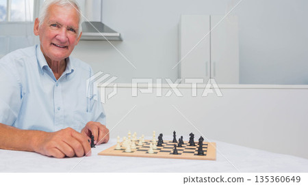 Senior man moving black chess piece on wooden chessboard in kitchen with white cabinets, copy space Senior man moving black chess piece on wooden chessboard in kitchen with white cabinets, copy space 135360649