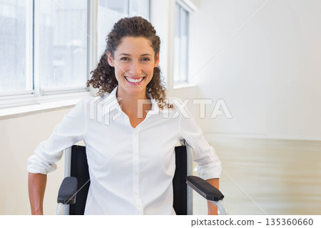 Female professional smiling while resting on wheelchair in bright corridor with large windows Female professional smiling while resting on wheelchair in bright corridor with large windows 135360660