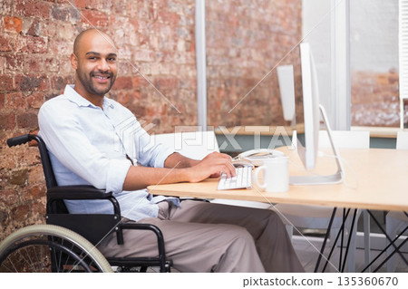 Mid adult man sitting in wheelchair typing on keyboard at desk in modern office, copy space 135360670