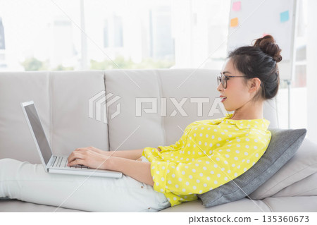 Chinese woman reclining on gray sofa typing on laptop near flip-chart in living room, copy space 135360673