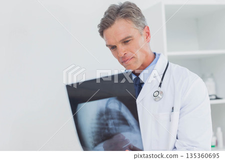Middle-aged male doctor wearing lab coat and holding chest X-ray film in exam room with stethoscope Middle-aged male doctor wearing lab coat and holding chest X-ray film in exam room with stethoscope 135360695