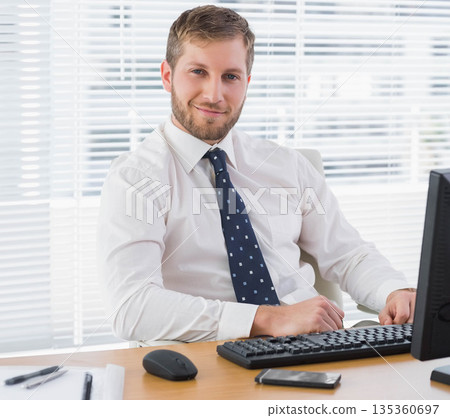 Man wearing white shirt and blue tie typing at office desk with keyboard, monitor and smartphone 135360697