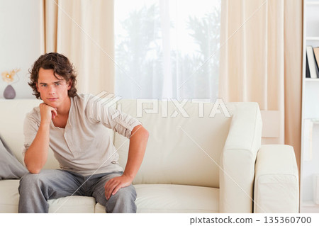 Man leaning, resting chin on hand on off-white sofa in sunlit living room, copy space 135360700